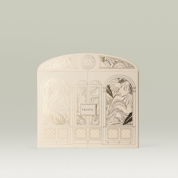 Tropic arch-shaped gift box in cream, featuring an illustrated building facade with gold foil tropical palm leaf detailing, arched windows, and a checkerboard base panel.